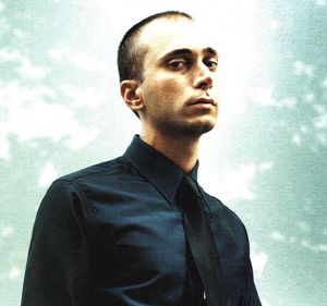 에디 슬리먼이 어두운 바지와 셔츠를 입고 카메라를 응시하는 모습.