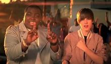 두 젊은 남자가 대화하고 있다. 왼쪽에 있는 남자는 손을 들고 검지 손가락으로 위를 가리키고 있으며 회색 재킷과 흰색 셔츠를 입고 있다. 오른쪽에 있는 남자는 왼팔을 가슴에 대고 있으며 빨간 셔츠와 검은색 티셔츠를 입고 있다. 그들 뒤에는 여러 사람이 손을 들고 있다.