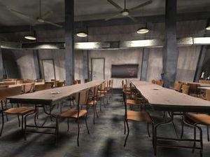 회색 벽에 두 개의 문이 있고, 중앙에 칠판이 있으며, 천장 선풍기와 매달린 조명이 머리 위에 있습니다. 전경에는 두 개의 긴 카페테리아 테이블이 배경으로 확장되어 있으며, 각 테이블에는 10개의 의자가 있습니다. 다른 테이블은 왼쪽과 오른쪽에 부분적으로 보입니다.