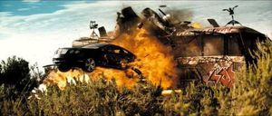 낙서로 덮인 버스 옆에서 검은색 차가 공중으로 날아오르는 모습, 버스가 폭발하고 있다.