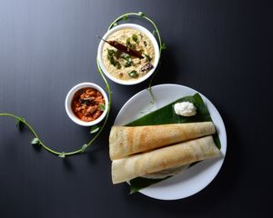 Two dosa rest next to a dollop of butter on a plantain leaf: The sauces have separate bowks.