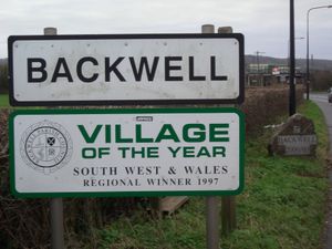 흰색 배경에 검은 글씨로 Backwell이라고 쓰여 있고, 그 아래에는 1997년 올해의 마을 사우스 웨스트 및 웨일스 지역 수상자라는 표지판이 있는 도로 표지판.