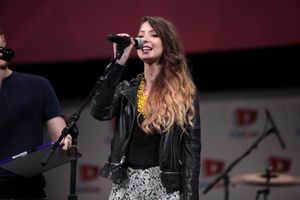 백인 여성으로 갈색 머리에 마이크에 대고 말하고 있다.