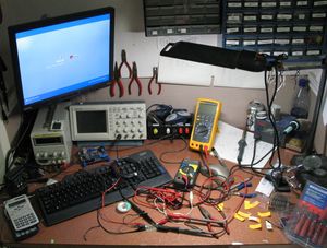 취미 전자 공학 애호가의 작업대. 오실로스코프, PC, 2개의 디지털 멀티미터, 납땜 스테이션, 포켓 계산기, 소형 공구, 안정화된 전원 공급 장치 및 전자 부품 보관함이 벽에 걸려 있다