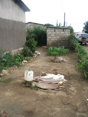 일반적인 문제: 우물 (앞쪽)이 재래식 화장실 (뒤쪽 벽돌 건물)과 매우 가까워 지하수 오염을 유발 (잠비아 루사카의 예시).