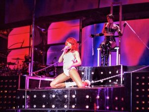 빨간 머리의 여성이 흰색 PVC 바디 수트를 입고 높은 플랫폼에서 공연하고 있다. 그녀는 마이크에 대고 노래를 부르고 있으며 무대에 수갑이 채워져 있다.