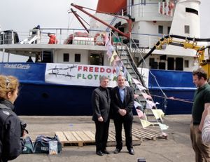 배 앞에 정장을 입은 두 남자. 배에는 <i>레이첼 코리</i>라는 이름이 적혀 있고 "자유 함대"라는 배너가 걸려 있다