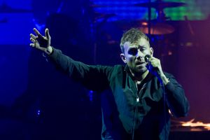 A middle-aged man standing in front of a coloured background with his right arm raised. He is holding a microphone in the opposite hand.