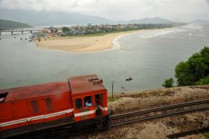 전경에는 붉은색 디젤 기관차가 랑꼬 해변과 바다를 배경으로 산을 오르고 있다. 배경에는 산들이 있다.