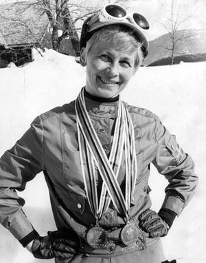 웃는 짧은 금발 여성은 장갑을 낀 트랙 수트를 입고, 흰색 스키 고글을 쓴 모자를 쓰고 있습니다. 그녀는 허리에 손을 얹고 목에 메달 두 개를 걸고 있습니다. 뒤에는 햇살이 비치는 눈 덮인 풍경이 펼쳐져 있습니다.