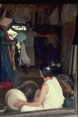 1971년 두린굿 의식. 주무당 (왼쪽)이 노래를 부르며 서 있고, 두 명의 견습 무당 (오른쪽 하단)이 북을 치며, 여성 환자 (가운데)가 춤을 춘다. 신들의 종이 그림이 천장에 걸려있다.