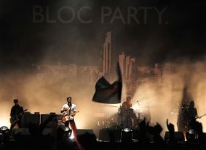 Two guitarists, a drummer, and a bassist are performing on a smoky stage lit by white stage lights. The stage background is a black tarpaulin emblazoned with "BLOC PARTY." and a large black-and-white cityscape below it. Most of the crowd have their hands up in the foreground, which also includes a waving flag.
