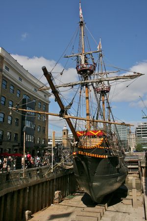 템스강의 작은 부두에 정박해 있는 골든 하인드 복제선의 앞모습. 주변의 높은 건물들과 함께 보인다.