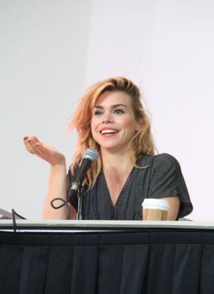 금발에 갈색 눈을 가진 20대 초반의 여성이 웃고 있으며, 흰색 티셔츠와 검은색 민소매 상의, 분홍색 스카프를 착용하고 있습니다.