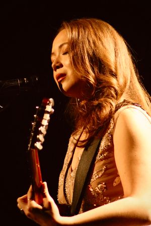 2024년 콘서트에서 Laufey의 측면 초상화. 그녀는 스포트라이트를 받으며, 어쿠스틱 기타를 연주하고 눈을 감고 서 있는 마이크에 대고 노래를 부르고 있다.