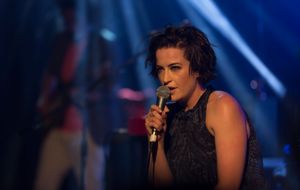 무대에서 공연하는 27세 여성이 상반신 샷으로 묘사되어 있습니다. 그녀는 왼쪽 측면을 바라보며 오른손에 든 마이크에 대고 노래합니다. 그녀는 땀에 젖은 검은 머리카락에 어두운 드레스를 입고 있습니다. 그녀의 손톱은 금색 반짝이로 칠해져 있습니다. 그녀 너머에는 장비와 동료 음악가들이 있지만 초점이 맞지 않아 흐릿하게 남아 있습니다.