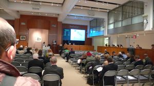 잠재 입찰자들이 접이식 금속 의자 줄에 앉아 있습니다. 프로젝터 스크린은 인터넷을 통해 들어오는 입찰을 표시하는 반면, 측면 벽을 따라 전화기를 든 사람들은 전화를 통해 전달된 입찰을 처리합니다. 대부분의 사람들은 고개를 숙이고 활력이 없어 보이며, 이 일련의 경매는 잠시 지속될 수 있습니다.