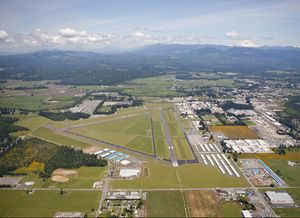 격납고, 창고, 열린 잔디밭으로 둘러싸인 세 개의 각진 활주로가 있는 공항. 배경에는 산과 삼림 지대가 보입니다.