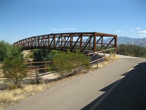 Photo shows a rust-colored truss footbridge with mountains in the right background.