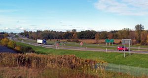 평탄한 지형을 가로지르는 분할 고속도로. 오른쪽에는 "마지막 미국 출구/Derniere Sortie EE. U."라고 적힌 영어 및 프랑스어 표지판이 있다. 왼쪽에는 도로가 카메라에서 멀어지고 약간의 언덕 위로 올라가 키가 큰 안테나가 있는 더 조밀한 지역으로 이어진다.