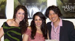 세 사람이 나란히 서서 카메라를 보고 미소를 짓고 있는 모습. 왼쪽과 중앙에는 20대 중반의 백인 여성 두 명이 있는데, 둘 다 긴 짙은 갈색 머리를 하고 있다. 왼쪽 여성은 검정색과 녹색 옷을 입고 있고, 중앙 여성은 분홍색 옷을 입고 있다. 오른쪽에는 30대 초반의 일본인 남자가 있는데, 머리 중앙에 가르마가 있고 어깨에 닿는 짙은 머리를 하고 있다. 그는 핑크색 오픈 칼라 셔츠와 검정색 재킷을 입고 있다.