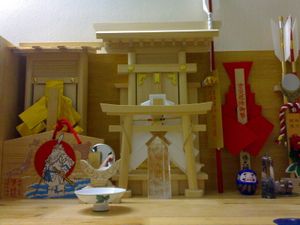 작은 소나무 신사로, 깃발로 장식된 평범한 도리이, 컵 앞의 작은 거울, 파란색 다루마 인형이 특징이다.